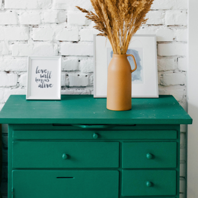 A green cabinet in fron of a white brick wall, and two images and a flower pot with grain on top of it.