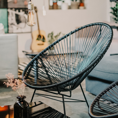 A black string chair in the middle of a living room. there  are some items in the foreground and the background.